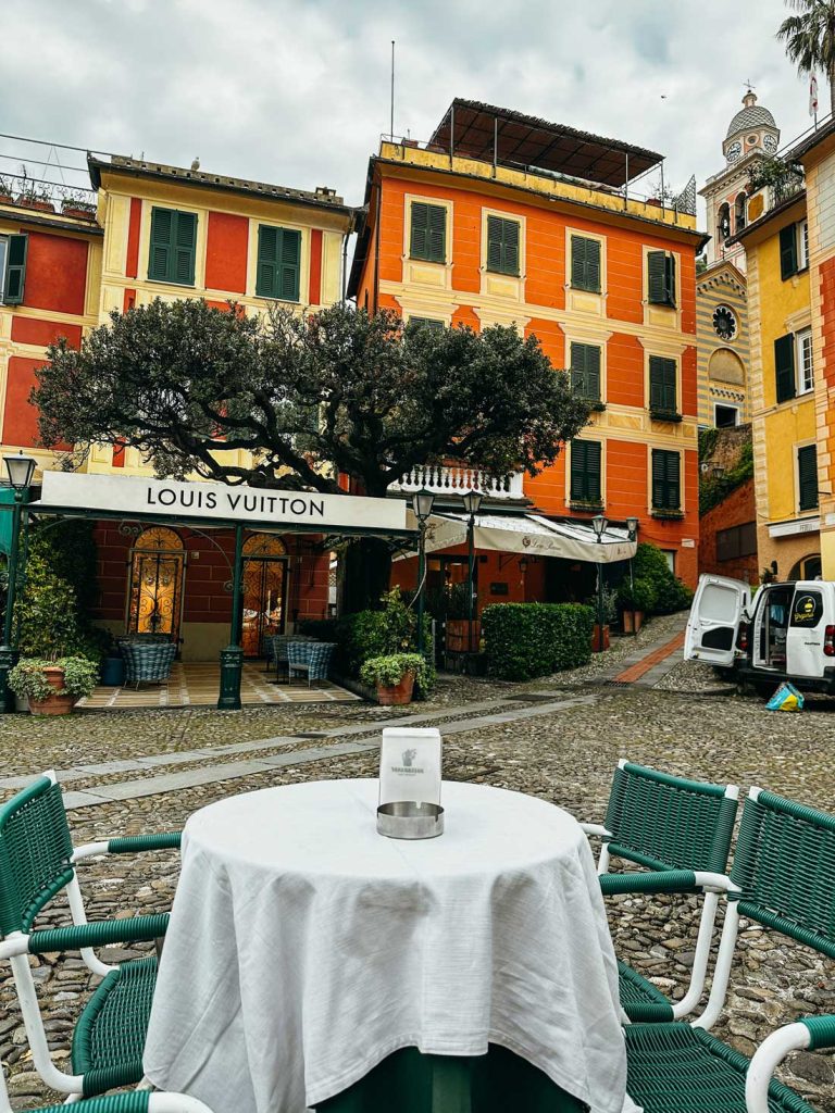 Portofino Or Cinque Terre: Which Is Better? 18 A cobblestone plaza in Portofino with a Louis Vuitton storefront framed by colorful orange and yellow buildings and a round table with green chairs in the foreground.