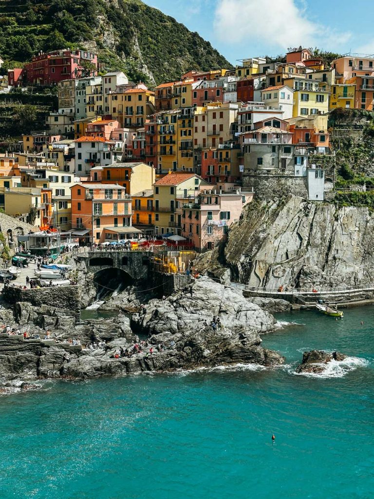 Portofino Or Cinque Terre: Which Is Better? 33 A scenic coastal shot of Manarola, Cinque Terre, with pastel homes stacked atop rocky cliffs above teal Mediterranean waters.
