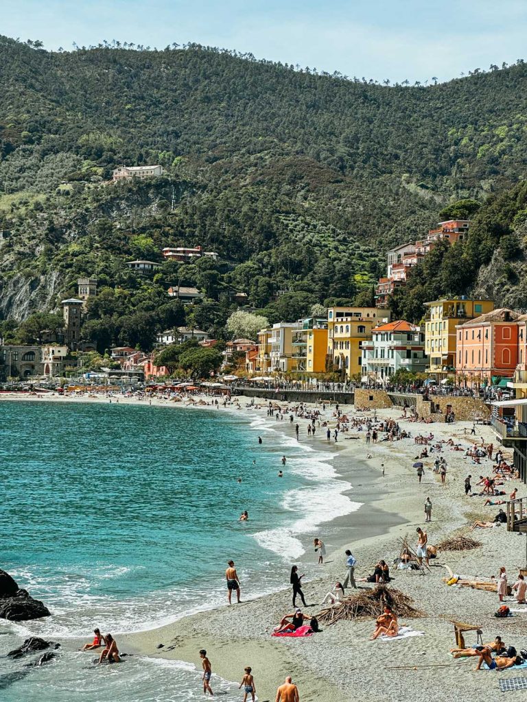 Portofino Or Cinque Terre: Which Is Better? 22 A sunny beach in Monterosso al Mare, Cinque Terre, filled with sunbathers and swimmers with a backdrop of lush hills and colorful buildings.