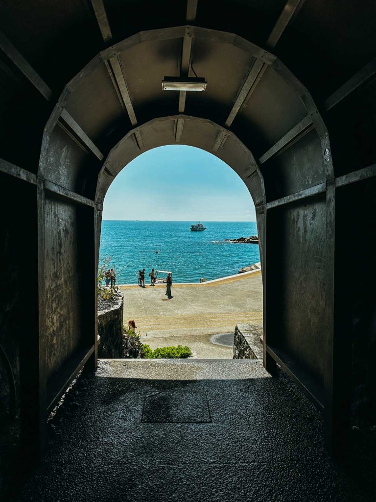 Portofino Or Cinque Terre: Which Is Better? 32 A stone tunnel opens to a bright coastal walkway and deep blue sea with a boat in the distance and people walking by the water.