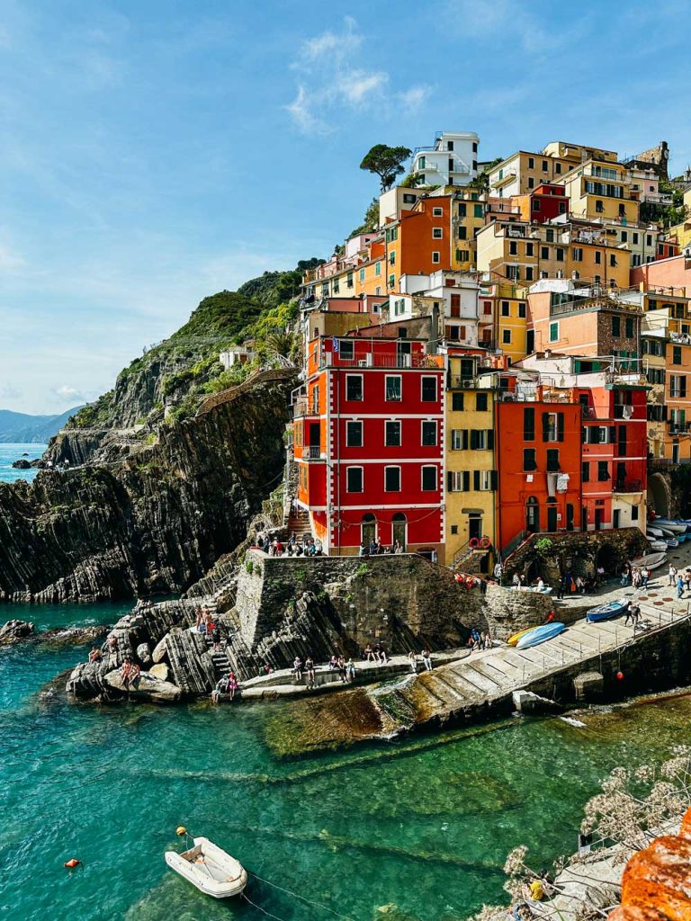 Portofino Or Cinque Terre: Which Is Better? 12 A vivid seaside scene in Riomaggiore featuring red and yellow cliffside buildings, tourists exploring the shoreline, and boats floating in calm green waters.