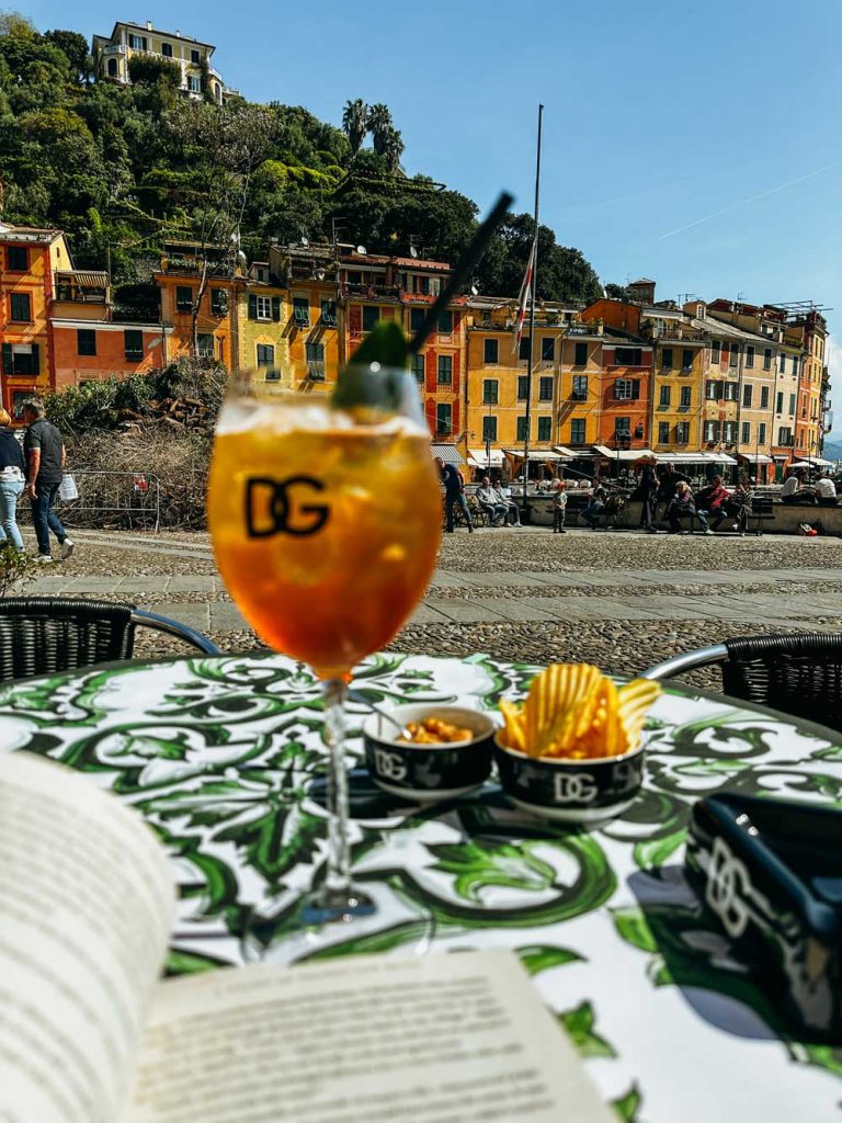 Portofino Or Cinque Terre: Which Is Better? 16 A cocktail and snacks are set on a Dolce & Gabbana café table with a scenic view of pastel buildings and a hillside in Portofino.