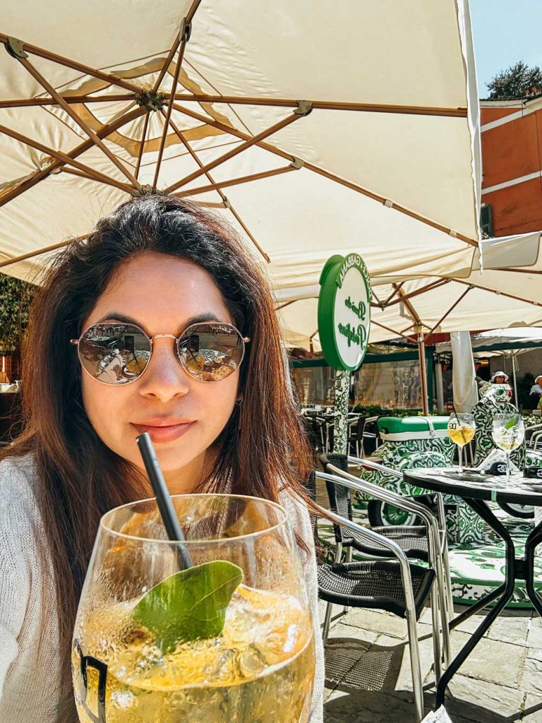 Portofino Or Cinque Terre: Which Is Better? 30 A woman in sunglasses sips a cocktail at an outdoor Dolce & Gabbana café under large white umbrellas with stylish green patterned chairs around her.