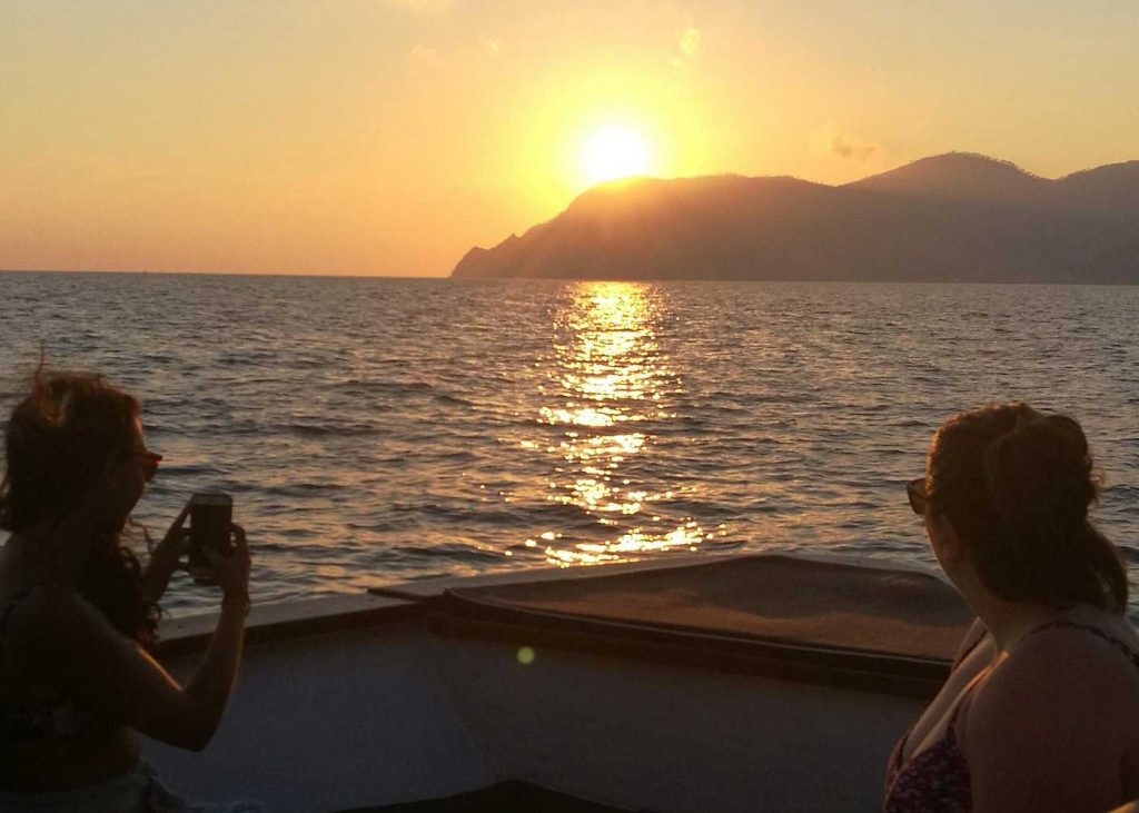 Portofino Or Cinque Terre: Which Is Better? 29 Two women sitting on a boat admire a glowing sunset over the sea with silhouetted mountains in the distance and golden reflections on the water.