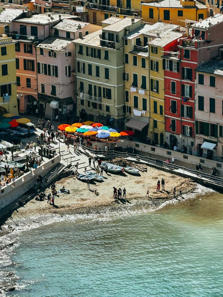 Portofino Or Cinque Terre: Which Is Better? 14 Close-up of the beachfront in Vernazza where people relax near colorful umbrellas and small boats in front of pastel buildings.