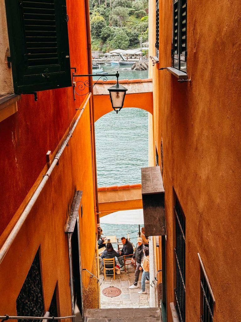 Portofino Or Cinque Terre: Which Is Better? 13 A narrow alleyway between orange buildings in Portofino opens to a waterfront café where people are dining with a view of the boats and sea.