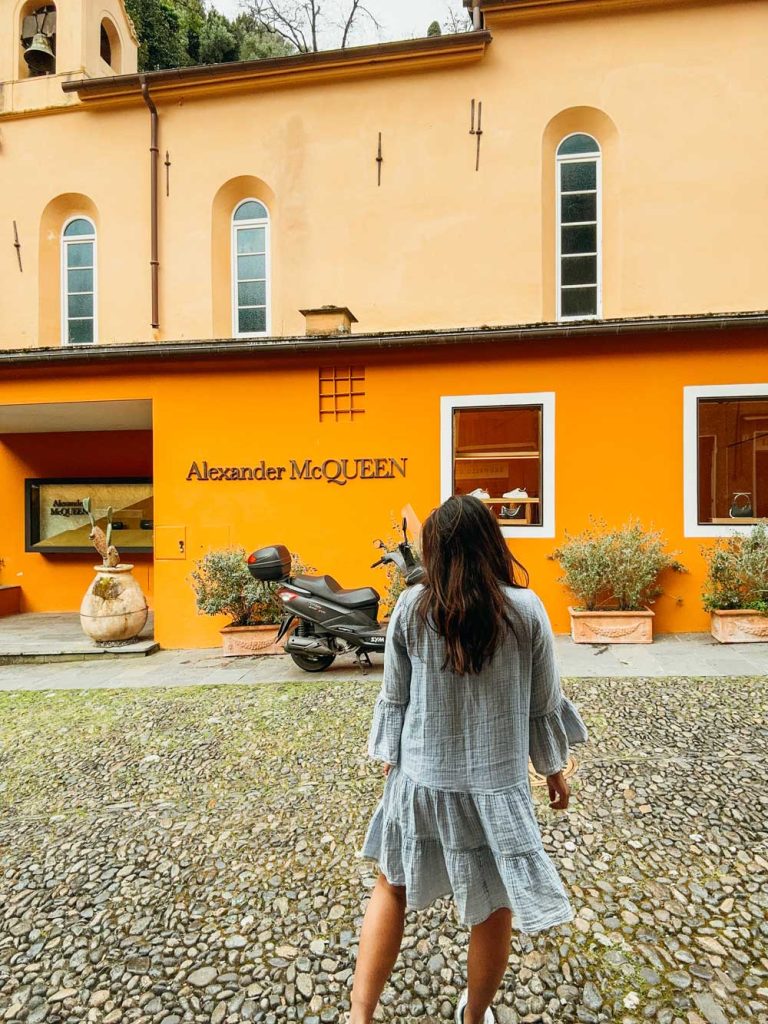 Portofino Or Cinque Terre: Which Is Better? 23 A woman in a light blue dress walks toward a bright orange Alexander McQueen storefront with cobblestone pavement and a motorbike parked nearby.