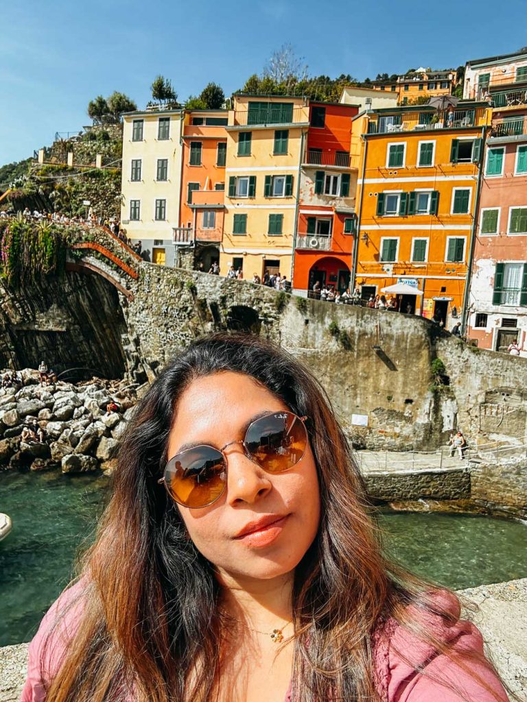 Portofino Or Cinque Terre: Which Is Better? 31 A woman in sunglasses takes a selfie with bright pastel buildings and a stone bridge in Riomaggiore, Cinque Terre in the background.