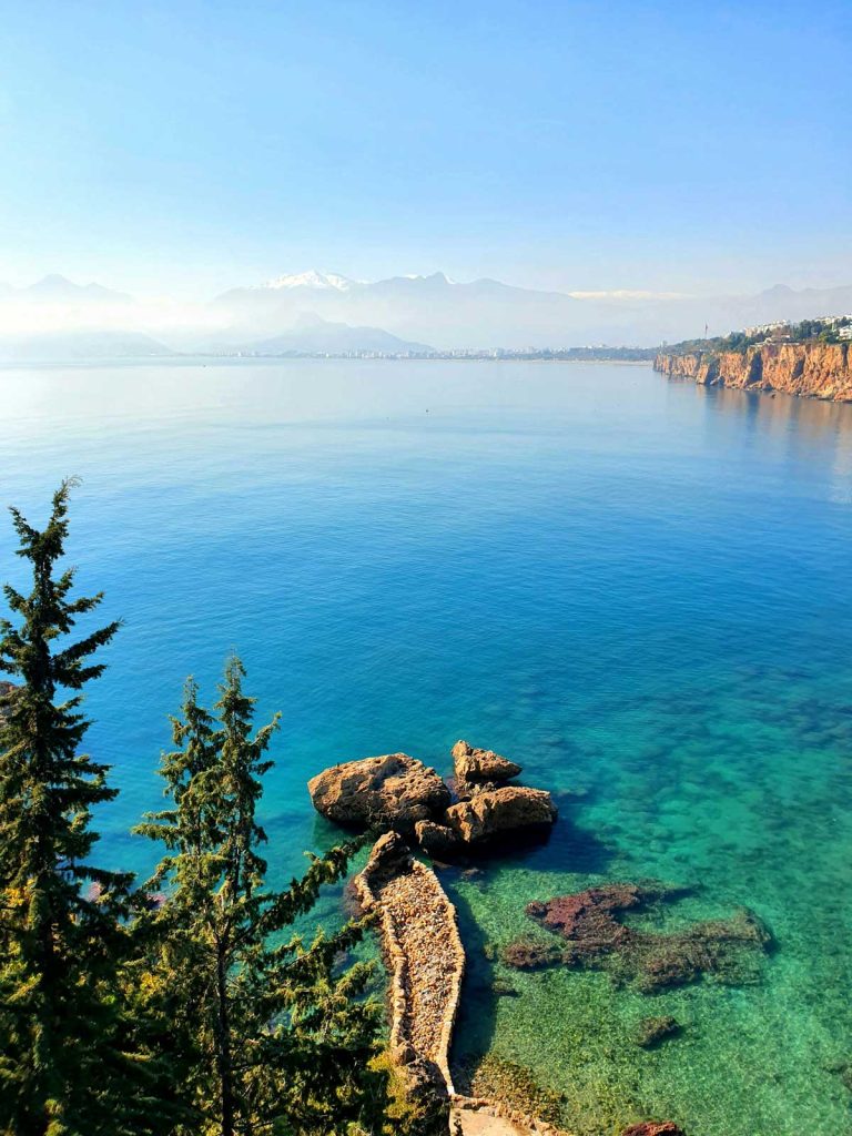 20 Best European Summer Destinations To Visit This Year 39 Crystal clear turquoise sea surrounds rocky cliffs with hazy mountains in the distance along a Mediterranean coastline. This serene coastal view captures the beauty of European summer destinations perfect for a relaxing seaside escape.