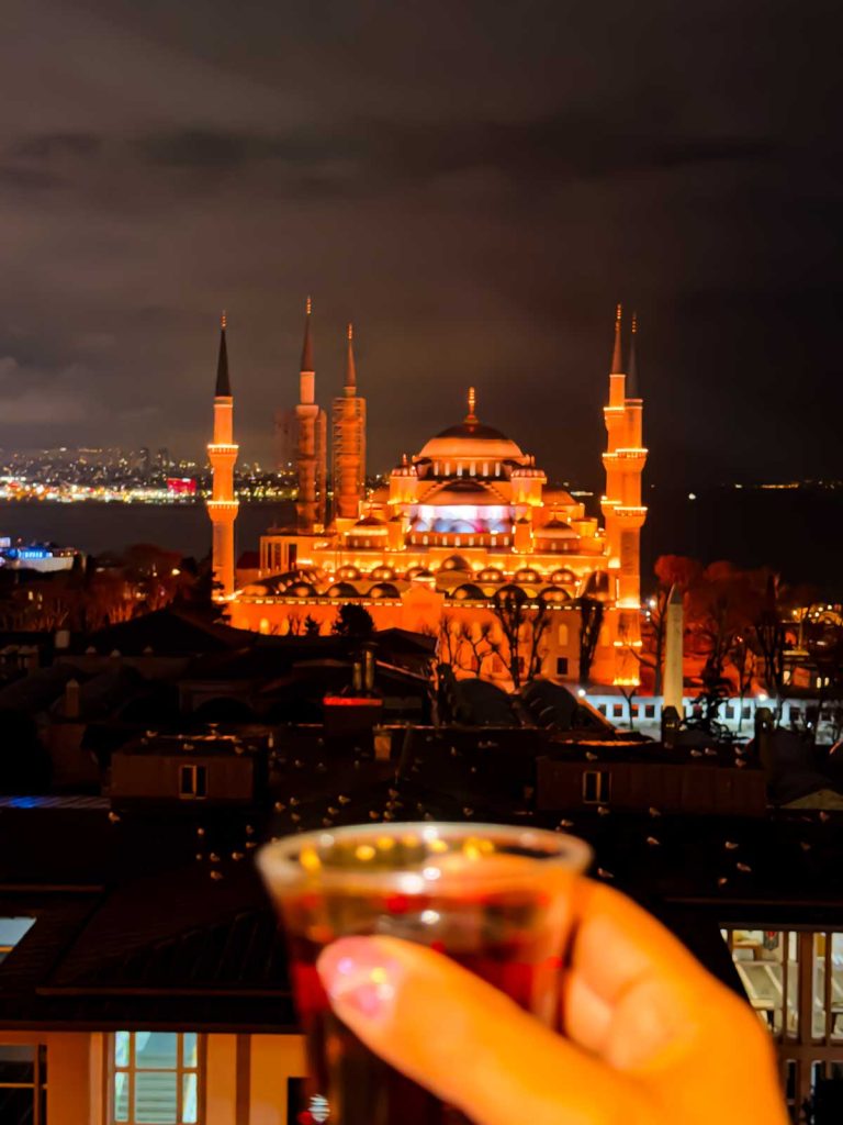 20 Best European Summer Destinations To Visit This Year 30 A glowing mosque with tall minarets lights up the night skyline as a hand holds a small glass of tea in the foreground. The illuminated cityscape captures a magical evening travel moment in Istanbul.