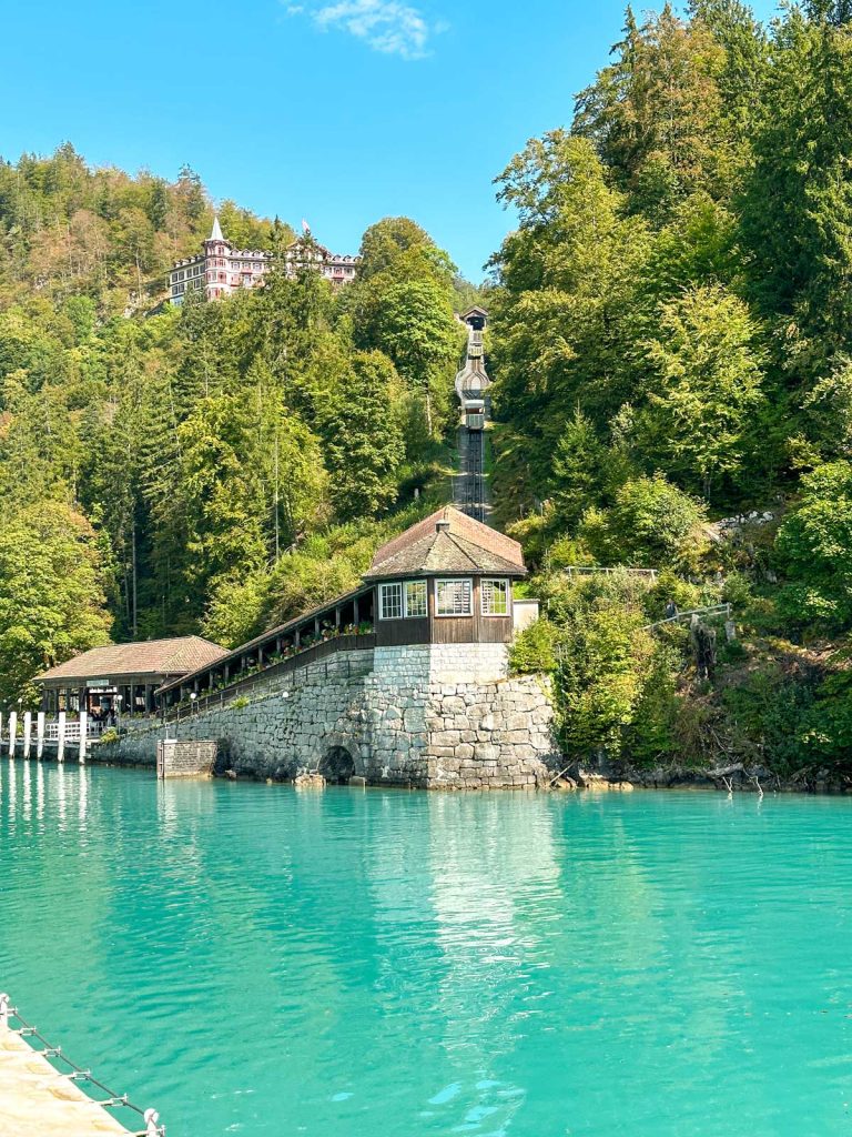 20 Best European Summer Destinations To Visit This Year 22 A turquoise lake sits beside a stone building with a steep hillside and forest rising behind it under a bright blue sky. The peaceful waterfront scene highlights scenic European summer destinations in the Alps.