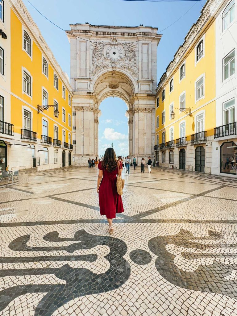 20 Best European Summer Destinations To Visit This Year 28 A woman in a red dress walks toward a monumental white arch framed by yellow buildings and patterned mosaic pavement. The sunny plaza scene captures the vibrant atmosphere of European summer destinations.