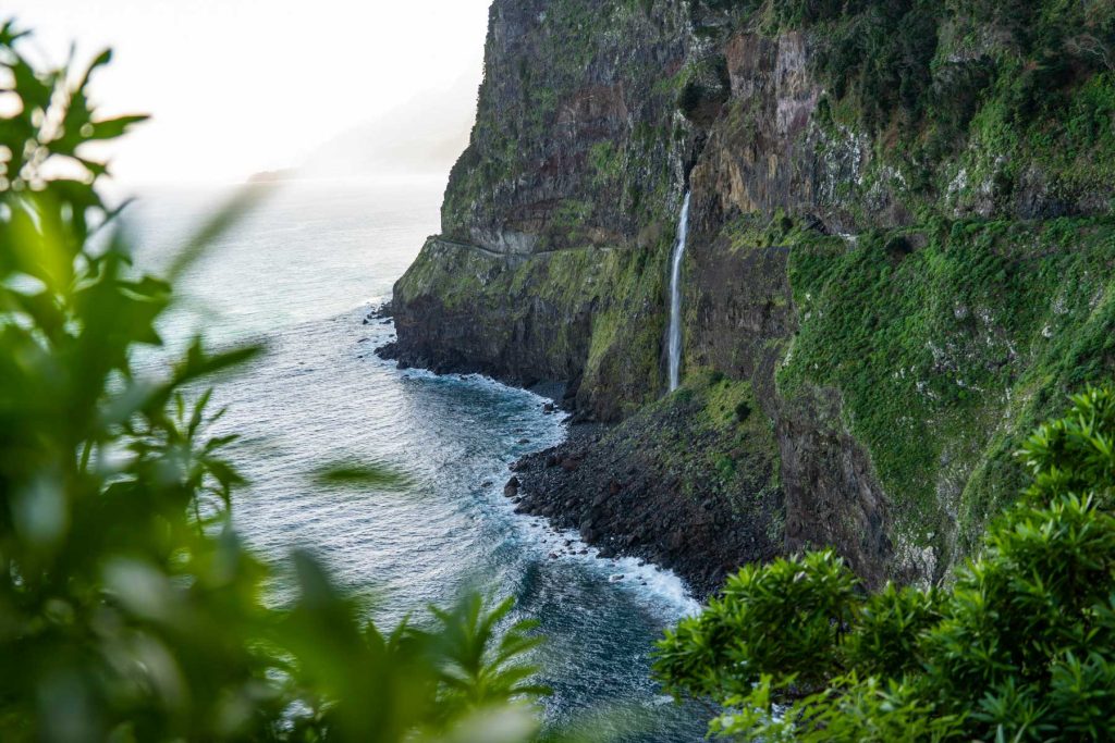20 Best European Summer Destinations To Visit This Year 37 A dramatic coastal cliff covered in lush greenery drops into the ocean with a thin waterfall cascading down to the rocky shore. The rugged landscape showcases the wild beauty of European summer destinations.