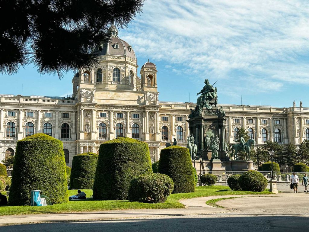 20 Best European Summer Destinations To Visit This Year 23 A grand historic building with a domed roof stands behind manicured green hedges and a large bronze statue in a city park. The elegant architecture represents cultural highlights often included on a Europe bucket list.