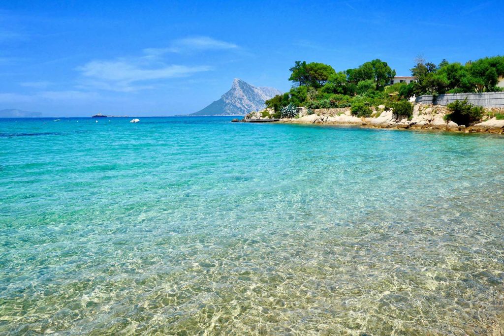 20 Best European Summer Destinations To Visit This Year 35 Crystal clear turquoise water stretches along a rocky shoreline with lush greenery and a mountain rising in the distance. The peaceful beach view reflects idyllic European summer travel along a scenic coastline.