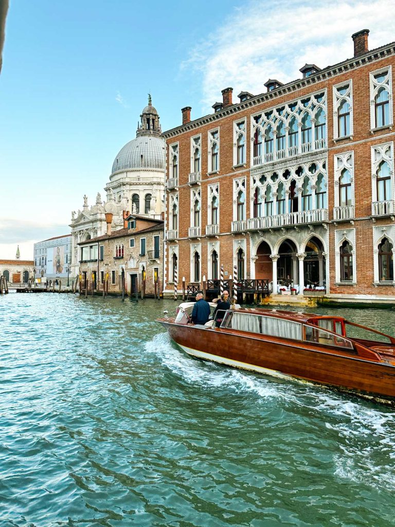 20 Best European Summer Destinations To Visit This Year 32 Golden sunset reflects across a Venetian canal as a gondolier rows past historic buildings and silhouetted domes. The glowing evening light captures a magical moment during a Venice itinerary.