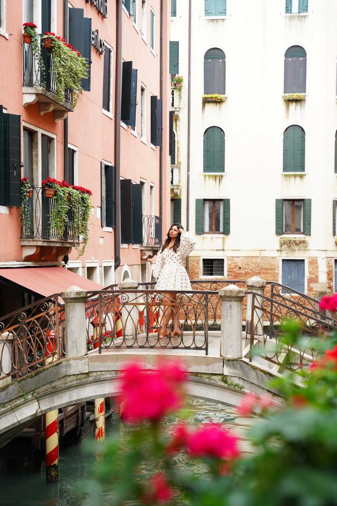 20 Best European Summer Destinations To Visit This Year 33 A woman in a white dress stands on a small arched bridge over a canal framed by pastel buildings with green shutters and flower boxes. The romantic city setting embodies classic European summer travel in a charming old town.