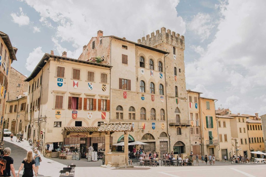 15 Amazing Day Trips From Florence, Italy 24 Sunlit Italian piazza lined with pale stone buildings and a medieval tower decorated with colorful shields. Small groups of people walk and gather in the square, showing the kind of historic town setting travelers may want to research carefully when learning how to plan travel abroad during uncertainty.