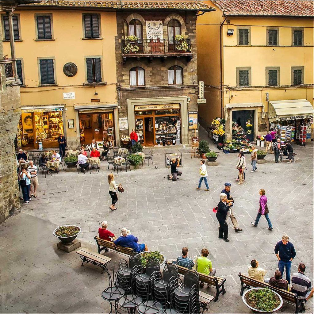 15 Amazing Day Trips From Florence, Italy 23 Busy town square with outdoor cafe tables, benches, and small storefronts beneath yellow buildings. Visible signs include "Toscana Servizi" on the balcony, "Enoteca Molteni dal 1937" above the central shop, "TABACCHI" on the right storefront, and "Piazza Repubblica" on the street sign, adding real local detail to the travel scene.