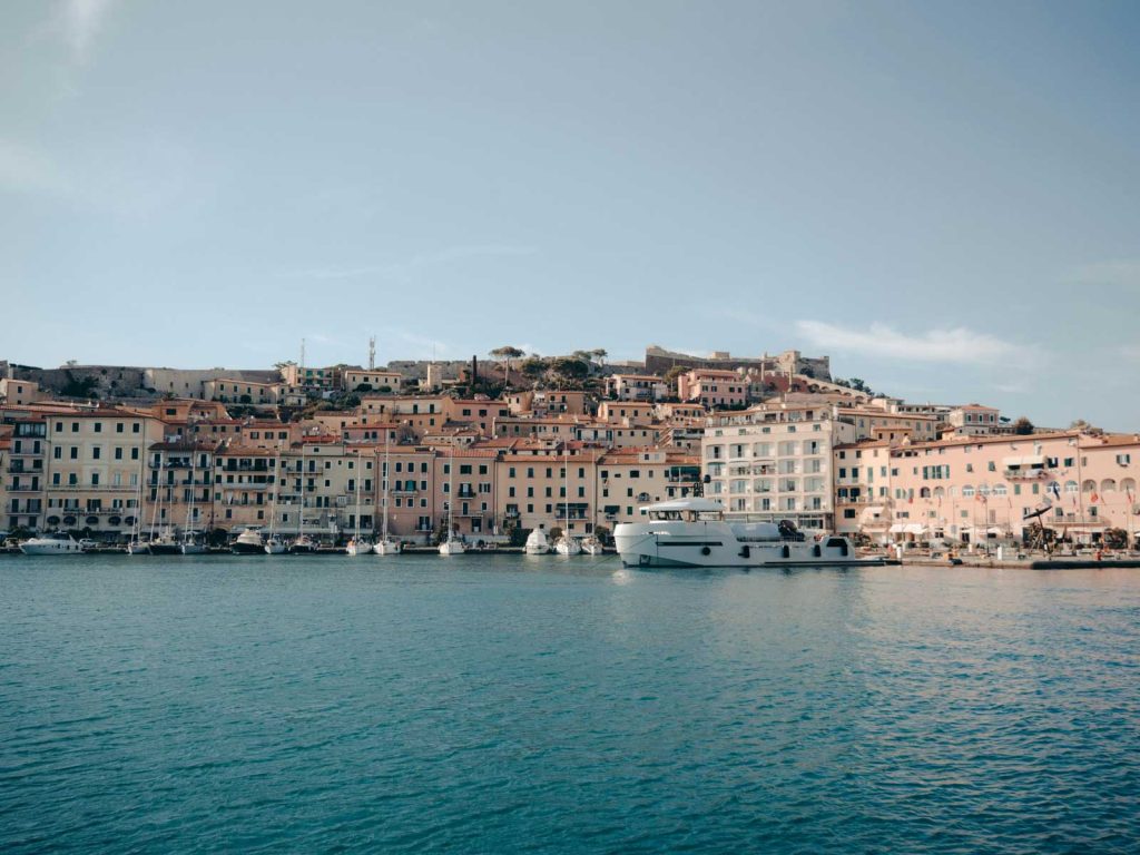 15 Amazing Day Trips From Florence, Italy 28 Waterfront town with pastel buildings stacked along the harbor and small boats moored on calm blue water. The coastal setting adds context for how to plan travel abroad during uncertainty because island and ferry based travel often requires flexible timing.