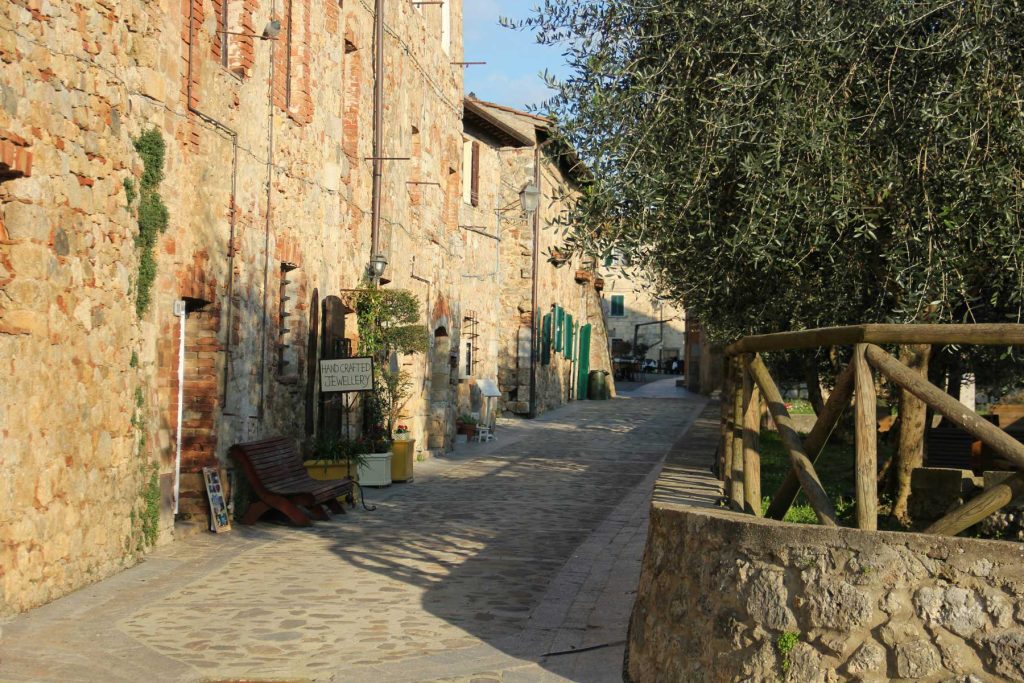 13 Best Towns In Tuscany 24 Narrow stone lane runs between old rustic buildings and olive trees in warm afternoon light. A small sign reading "HANDCRAFTED JEWELLERY" marks a quiet village street that suggests slower local travel and the need for adaptable plans.