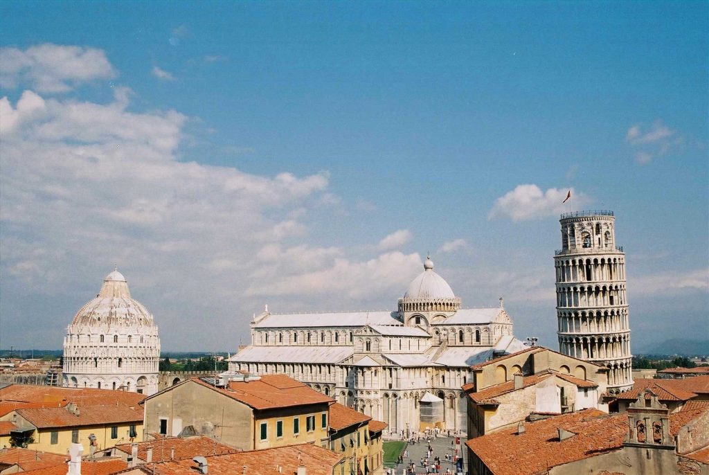 15 Amazing Day Trips From Florence, Italy 22 Wide view of Pisa with terracotta rooftops in the foreground and the cathedral complex rising in bright white stone. The Leaning Tower stands to the right against a blue sky, grounding the article in a recognizable European destination.