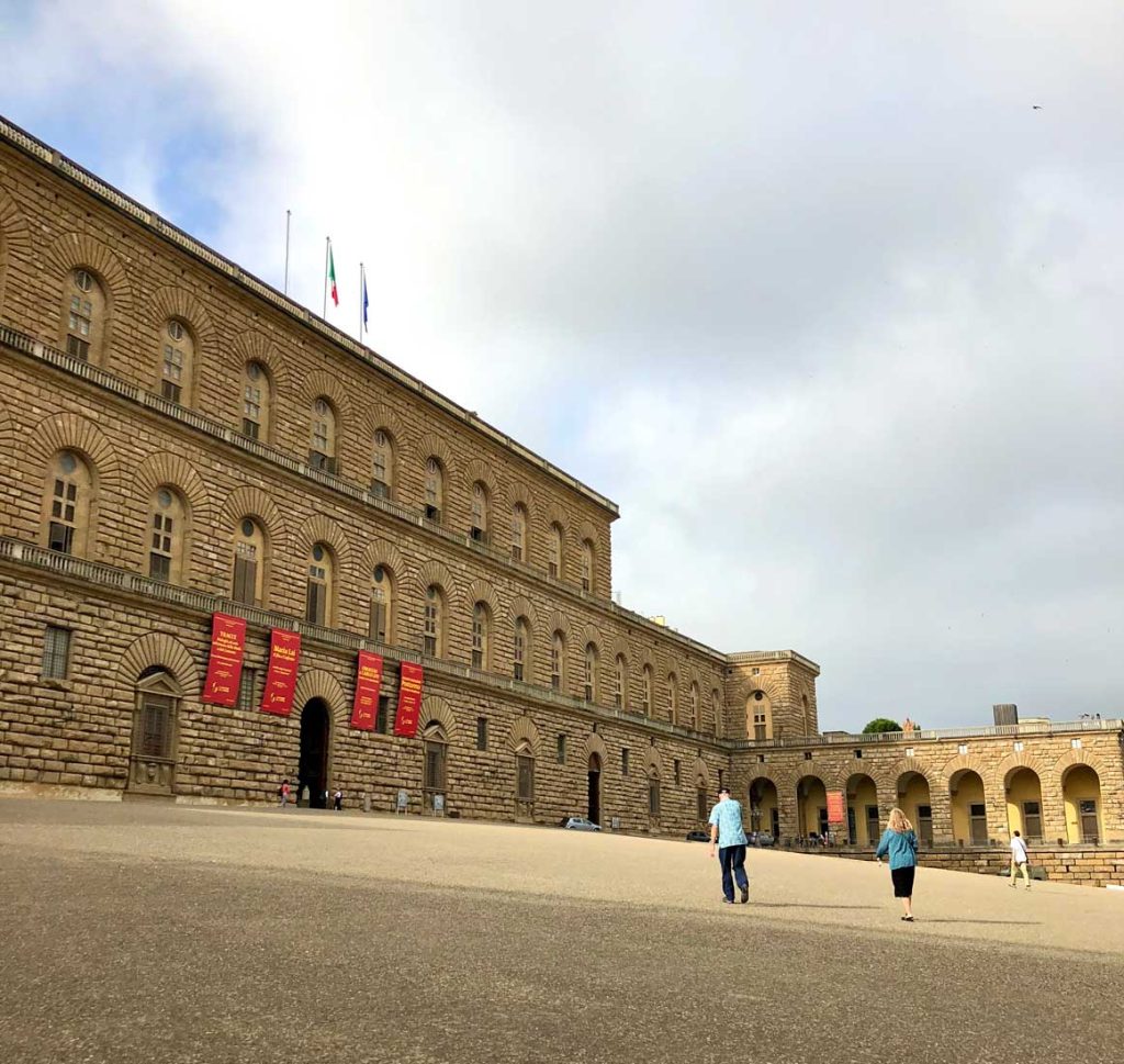 The Perfect 3 Day Florence Itinerary (From an Italy Travel Expert) 18 Wide courtyard outside Palazzo Pitti in Florence with a few visitors walking toward the large stone palace. Red museum banners hang near the arched entrance.