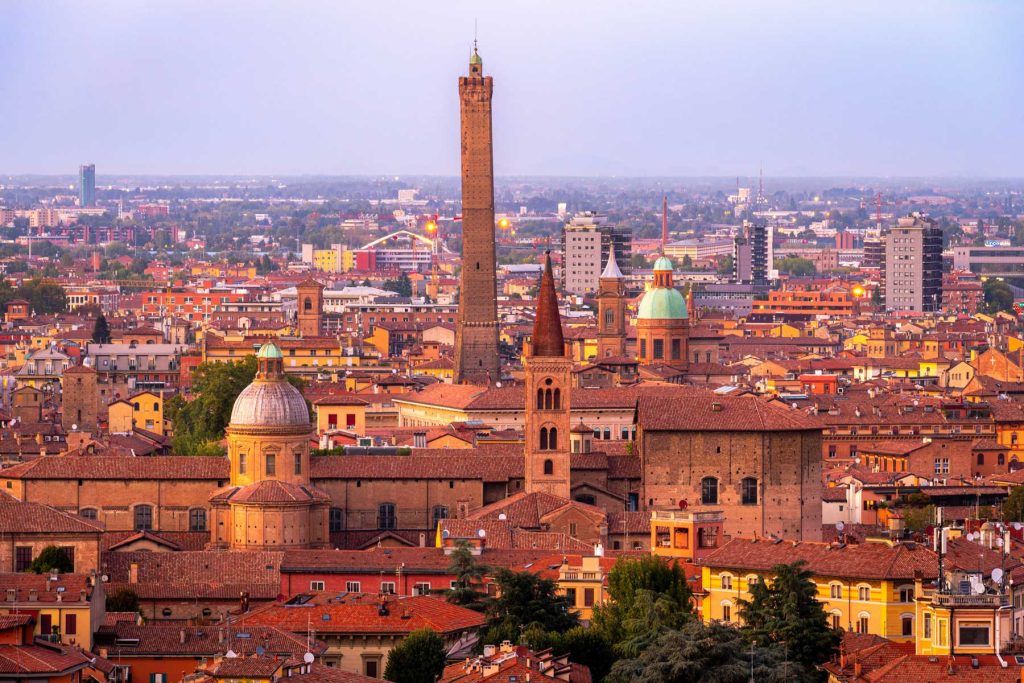15 Amazing Day Trips From Florence, Italy 26 Bologna skyline at dusk with the Two Towers rising above red rooftops and domed churches. Warm city lights and a purple evening sky frame the historic center.