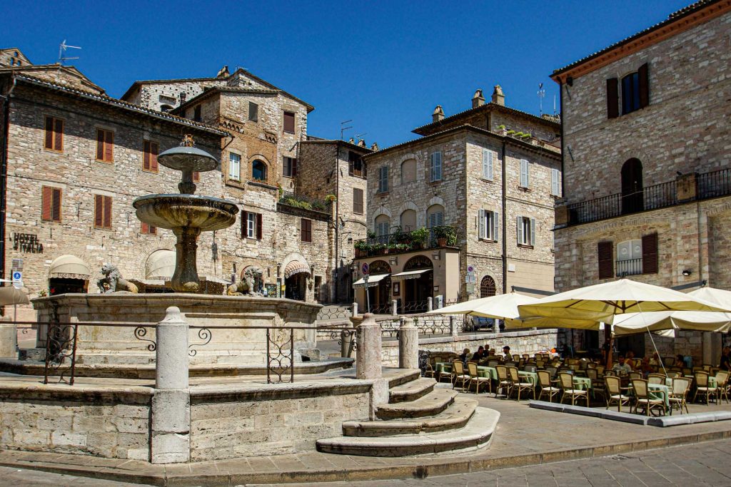 15 Amazing Day Trips From Florence, Italy 29 Historic stone piazza with a tiered fountain. Outdoor cafe tables sit beneath cream umbrellas beside medieval buildings.